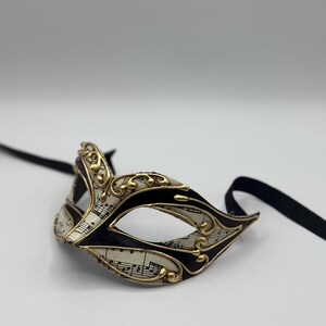 May include: A black and gold masquerade mask with a white background featuring musical notes. The mask has a black ribbon attached.