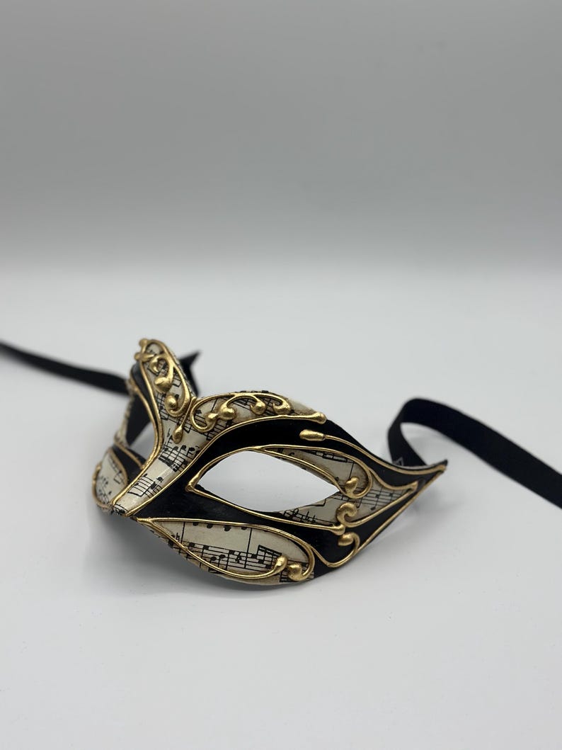 May include: A black and gold masquerade mask with a white background featuring musical notes. The mask has a black ribbon attached.