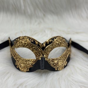 Pode incluir: Uma máscara de baile de máscaras preta e dourada com detalhes intrincados e ornamentados. A máscara apresenta uma base preta com detalhes em filigrana dourada e fitas pretas para amarrar. O design é simétrico e elegante, adequado para eventos formais.
