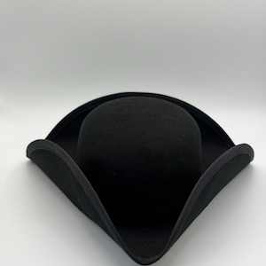 Puede incluir: Un sombrero tricornio negro con una corona redondeada y un ala ancha y levantada. El sombrero está hecho de un material similar al fieltro y se coloca sobre un fondo blanco. Un sombrero de estilo clásico.