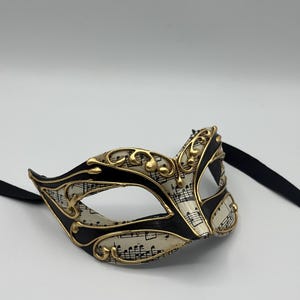 May include: A black and gold masquerade mask with a white background featuring musical notes. The mask has a black ribbon attached.