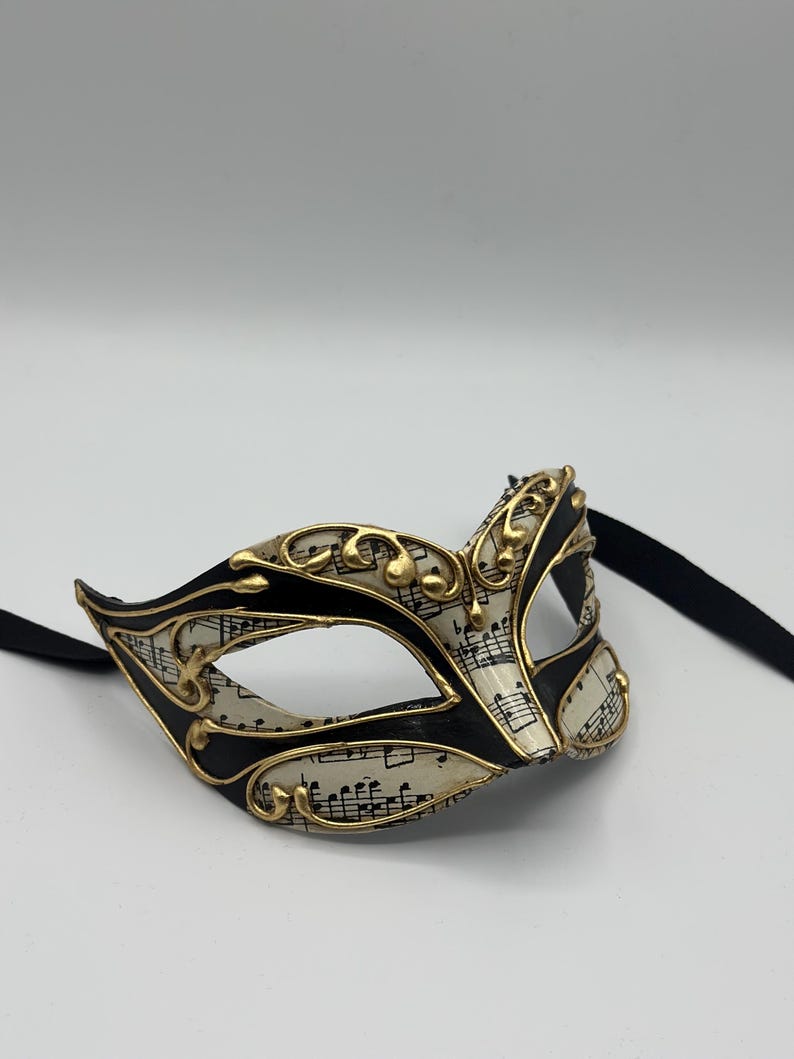 May include: A black and gold masquerade mask with a white background featuring musical notes. The mask has a black ribbon attached.