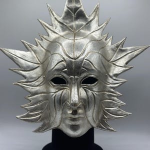 Pode incluir: Máscara prateada com design solar. A máscara apresenta um rosto com expressão serena, rodeado por elementos em forma de folha. A máscara é exibida em um suporte preto sobre um fundo cinza.