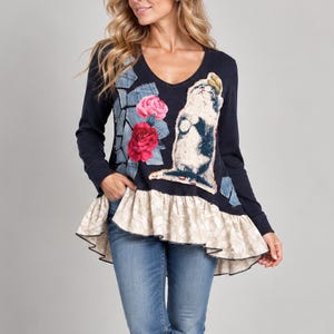 Könnte beinhalten: Eine langärmelige marineblaue Tunika mit V-Ausschnitt, einem gerüschten beigen Saum mit Muster und einem Grafikdruck auf der Vorderseite, der eine Katze, rote Rosen und Jeans-Patches zeigt, kombiniert mit einer blauen Jeans und braunen Lederstiefeletten.