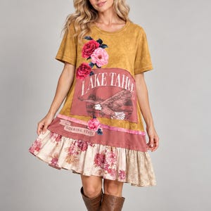 Puede incluir: Un vestido de manga corta color amarillo mostaza con un gráfico de un águila y el texto LAKE TAHOE y SOARING STATE. El vestido tiene un panel rectangular granate con detalles florales rosas y un dobladillo con volantes y estampado floral.