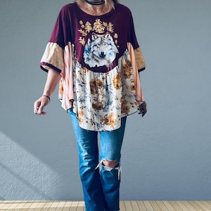 May include: A loose-fitting top featuring a maroon upper section with a central wolf graphic surrounded by gold pine trees. The lower portion is a cream-coloured fabric with a brown floral pattern and striped side panels, paired with blue jeans.