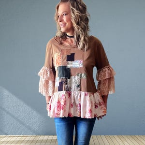 May include: A brown long-sleeve top featuring a patchwork design on the chest with various fabric textures and colors. The garment includes tiered lace sleeves and a ruffled hemline with a pink and white floral pattern, paired with blue jeans.