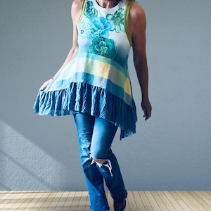 May include: A sleeveless tunic top featuring a white bodice with teal and green floral patterns, a mid-section with yellow and blue plaid horizontal stripes, and a ruffled blue hem. The top is paired with blue denim jeans that have a distressed knee.