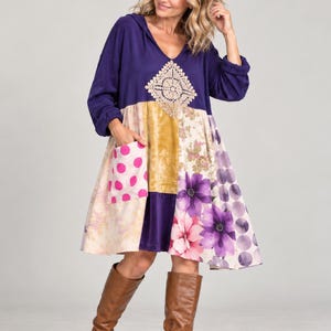 Puede incluir: Un vestido de manga larga con capucha que presenta un corpiño morado oscuro con una aplicación de crochet color crema en forma de diamante. La falda es un patchwork de estampados florales, lunares rosas y paneles morados, con botas de cuero.