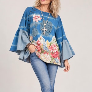 Puede incluir: Una blusa azul de manga larga con patrón tie-dye, mangas acampanadas anchas y un estampado floral en la parte inferior. La prenda incluye aplicaciones circulares tipo encaje en el pecho y un collar con colgante de cruz en cadena de cuentas.