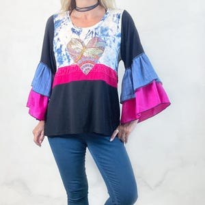 May include: A long-sleeved top with a heart-shaped design. The top features a tie-dye pattern, a pink stripe, and layered bell sleeves in blue and pink. The model is wearing blue jeans and brown boots.