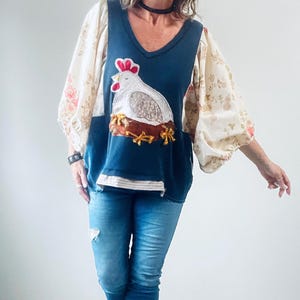 May include: A navy blue V-neck top with a white hen applique on the front. The top has floral print sleeves and is paired with blue jeans. The hen is sitting on a nest with gold-colored accents.