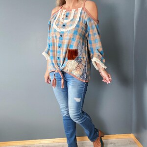 May include: A blue, tan, and brown plaid open-shoulder top with a knotted front and decorative patches. The top is worn with blue jeans and brown patterned boots. The outfit has a bohemian, country-western aesthetic.