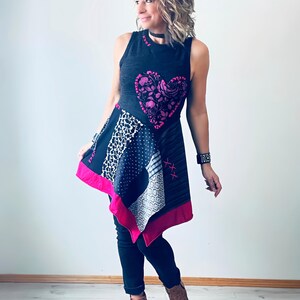 Puede incluir: Un vestido asimétrico sin mangas con un corpiño gris oscuro y una falda de retazos. El vestido presenta un gran diseño de corazón rosa y un ribete rosa brillante. La modelo lleva pantalones negros y botas marrones.