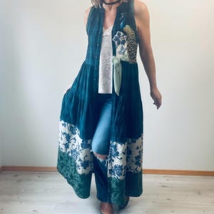 May include: A sleeveless, floor-length teal duster with a floral pattern on the bottom and a peacock applique. The duster is layered over a white top and blue jeans. A green ribbon is tied at the waist.