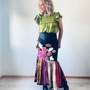 Puede incluir: Una mujer con un top verde con volantes y una falda negra con diseño floral. La falda tiene un dobladillo de patchwork con varios colores y patrones. También lleva botas negras con plataforma.