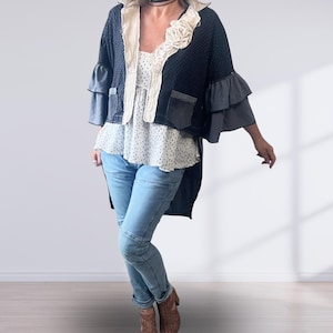 May include: A black jacket with a subtle dot pattern, featuring a cream-coloured ruffled collar, tiered bell sleeves in grey, and two front pockets. It is worn over a white sleeveless top with a small black dot pattern and light blue denim jeans.