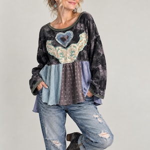 Könnte beinhalten: Ein langärmeliges Oberteil mit schwarz-grauem Batikmuster, einem zentralen Herzmotiv und einem dekorativen Rankenmuster. Der untere Bereich besteht aus einem mehrteiligen Patchwork-Saum in Blau-, Grau- und Dunkelbrauntönen mit Kreuzmuster.