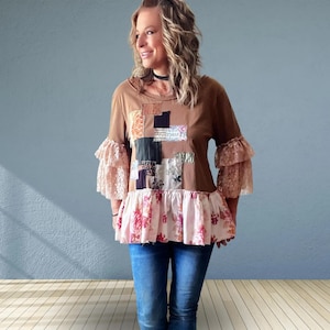 May include: A brown long-sleeve top featuring a patchwork design on the chest, double-layered lace ruffles on the sleeves, and a light pink floral-patterned ruffled hemline, paired with blue denim jeans and a black choker necklace.