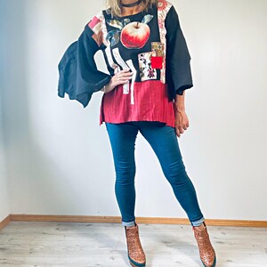 May include: A black top with an apple design, red and white accents, and wide sleeves. The top is paired with blue jeans and brown boots. The outfit is a unique combination of colours and textures.