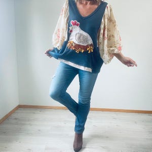 May include: A navy blue V-neck top with a white hen and nest applique. The top has floral print sleeves and is paired with light blue jeans. The hen applique features a red comb and a brown nest.