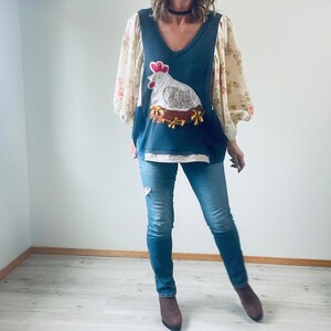 May include: A dark blue V-neck top with a large chicken applique on the front. The chicken is white and gray with a red comb, sitting on a brown nest. The top has floral print sleeves and is paired with blue jeans.