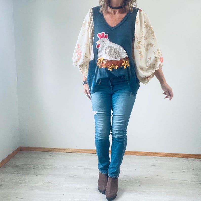 May include: A dark blue V-neck top with a white hen and nest applique. The top has floral patterned sleeves and is paired with blue jeans. The outfit is completed with brown ankle boots.