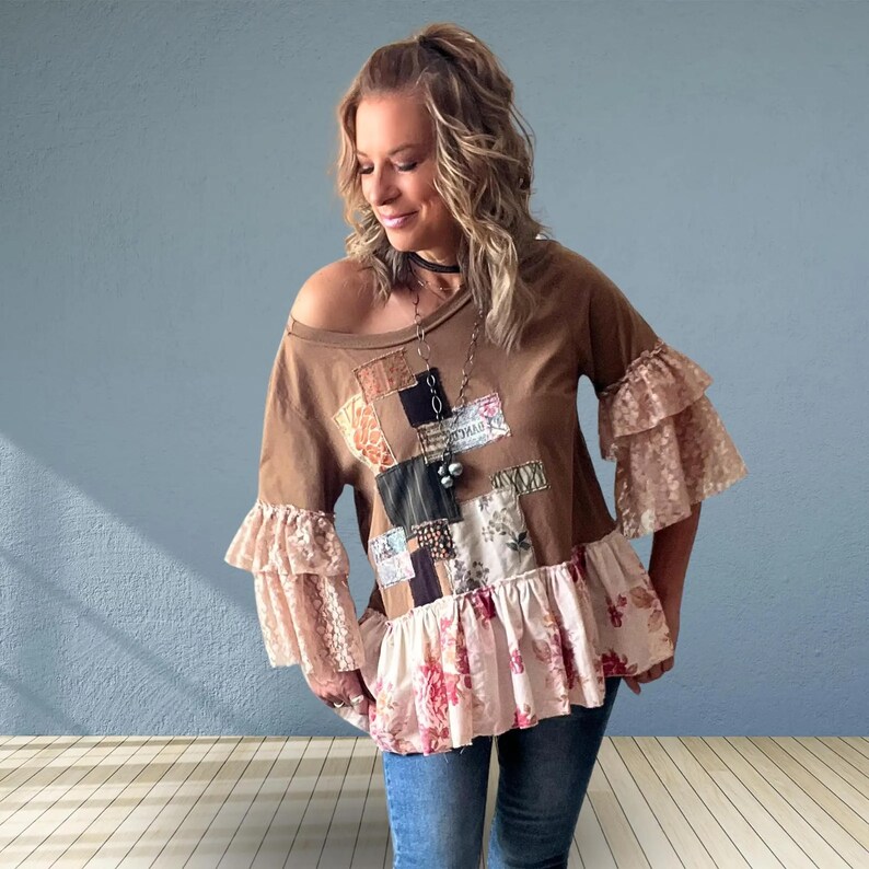 May include: A brown long-sleeve top featuring a patchwork design on the front with various fabric patterns, including floral and pinstripe prints. The garment has tiered lace ruffles on the sleeves and a ruffled floral hemline at the bottom edge.