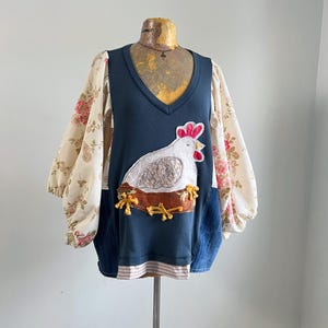May include: A unique, handmade top with a navy blue bodice and floral print sleeves. The front features a white chicken applique on a brown nest. The sleeves are a cream color with pink and red roses. The top has a V-neck and a striped hem.