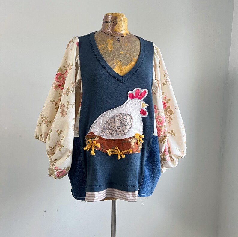 May include: A unique, handmade top with a navy blue bodice and floral print sleeves. The front features a white chicken applique on a brown nest. The sleeves are a cream color with pink and red roses. The top has a V-neck and a striped hem.