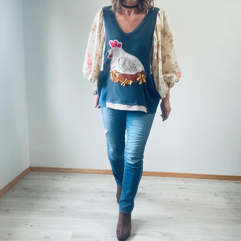 May include: A denim blue V-neck top with a chicken applique, paired with light wash blue jeans. The top has floral print sleeves. The chicken applique features a white body, red comb, and brown nest. The jeans have distressed details.