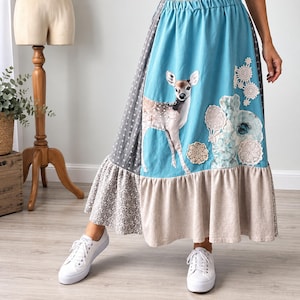 May include: A long, multi-paneled skirt featuring a bright blue section with a printed fawn graphic and white lace floral appliqués. The skirt includes side panels in grey with a white cross pattern and a grey damask print, finished with a beige hem.