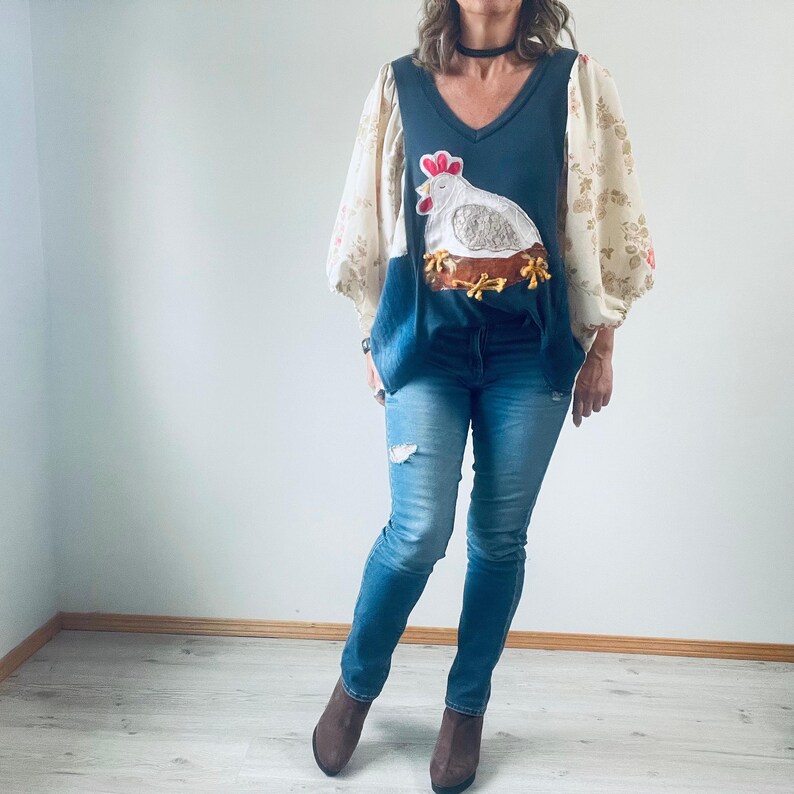 May include: A dark blue V-neck top with a chicken applique, paired with light blue jeans. The top features floral patterned sleeves. The chicken applique is white with a red comb and is sitting on a brown nest.