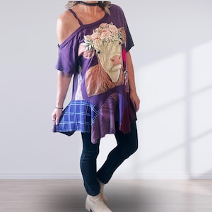 May include: A purple short-sleeved tunic featuring a graphic of a cow wearing a floral crown. The garment has an asymmetrical cold-shoulder neckline with a thin strap and a hemline panel with a blue and white grid pattern and purple fabric.
