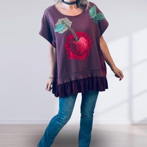 May include: A dark maroon short-sleeved top featuring a large cherry appliqué on the front. The cherry is rendered in shades of red with green fabric leaves and a textured lace-like background. The garment has a ruffled hemline at the bottom.