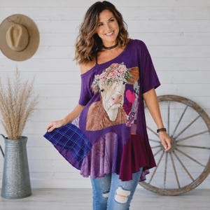 Może przedstawiać: Fioletowa tunika z krótkim rękawem z grafiką krowy w wianku z kwiatów. Odzież ma wzór w stylu patchwork z różnymi fioletowymi i niebieskimi motywami, w tym sekcjami w kratę i gładkimi, oraz luźny, asymetryczny dół.