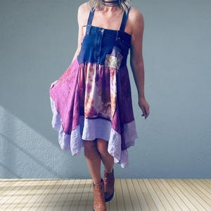May include: A sleeveless dress featuring a dark blue denim bodice with a button-down front and a chest pocket. The skirt is constructed from multiple panels of fabric in shades of magenta, purple, and lavender with a tie-dye and floral pattern.