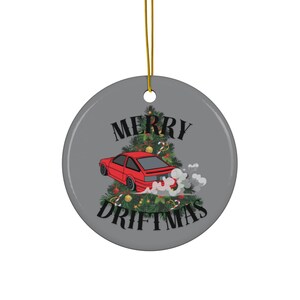May include: A round, gray Christmas ornament with a red car drifting in front of a Christmas tree. The text "MERRY DRIFTMAS" is printed on the ornament in black. The ornament hangs from a gold string.
