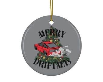 May include: A circular, grey Christmas tree decoration featuring a red car drifting in front of a Christmas tree. The words "MERRY DRIFTMAS" are printed in black. It hangs from a gold string.