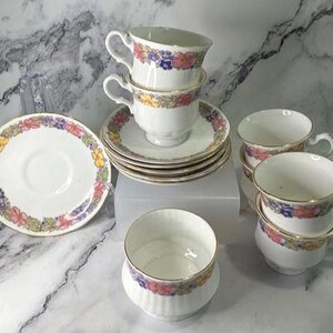 May include: A set of white porcelain teacups and saucers featuring a colourful floral pattern of pink, yellow, and blue flowers with green leaves. Each piece is finished with a thin gold rim. The set includes stacked cups and saucers.