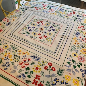 May include: A cream-coloured tablecloth with a vibrant floral design. The pattern includes red, yellow, and blue flowers with green foliage. The tablecloth has a central square design and a floral border.