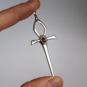 Könnte beinhalten: Ein silberner Anhänger in Form eines Ankh, einem Symbol des Lebens. Der Anhänger hat einen kleinen, oval geformten, roten Edelstein in der Mitte des Kreuzes. Der Ankh wird von einem Finger gehalten.