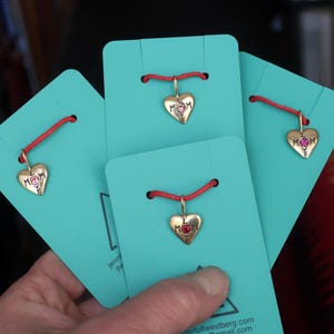 May include: Gold-colored heart-shaped pendants with the word "MOM" engraved on them, strung on red cords, displayed on turquoise cards. Some pendants have a pink gemstone. The cards have text and are held by a hand.