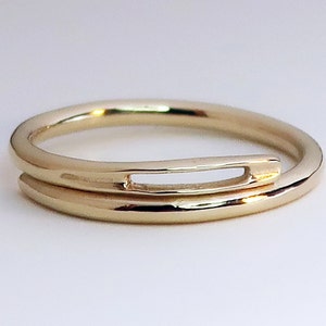 Puede incluir: Un anillo de oro con un diseño único que presenta una banda fina con una banda más gruesa envuelta alrededor, creando una forma recortada.