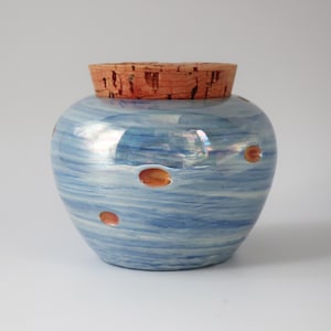 May include: A ceramic jar with a light blue, swirled pattern and small orange spots. The jar is topped with a cork stopper. The jar has a round shape and is likely used for storage or decoration.