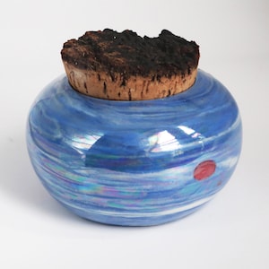 May include: A round, blue ceramic jar with a cork stopper. The jar has a glossy finish with swirling blue and white patterns, and a small red dot. The cork stopper is dark brown and textured.