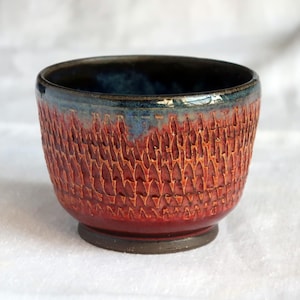 May include: A handmade ceramic bowl with a textured, carved exterior. The bowl features a gradient of colors, with a deep red base that transitions to a lighter red and then to a blue rim. The interior is a dark blue color.