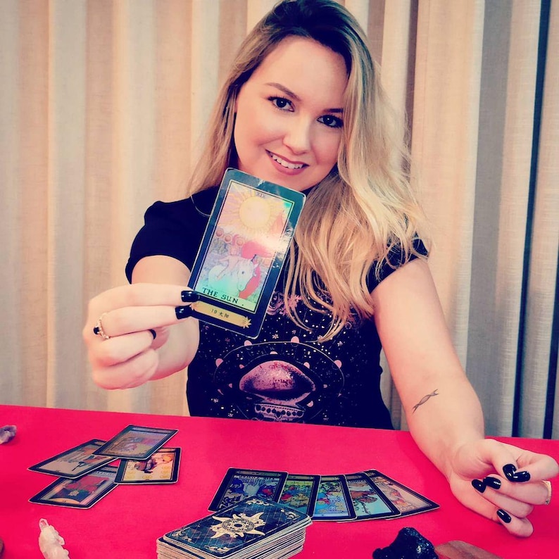 May include: A woman holds up a tarot card with the image of the sun and the text "THE SUN" and "XIX" on it. The card is surrounded by other tarot cards on a red table.