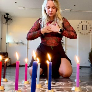 May include: A woman in a red floral top and black skirt is kneeling in front of a circle of lit candles. The candles are in different colors, including pink, purple, blue, and orange. The candles are on a white surface with a chalk drawing.