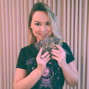 May include: A woman with blonde hair is holding a spread of tarot cards in her hands. The cards are decorated with intricate designs and symbols. The woman is wearing a black t-shirt with a celestial design.